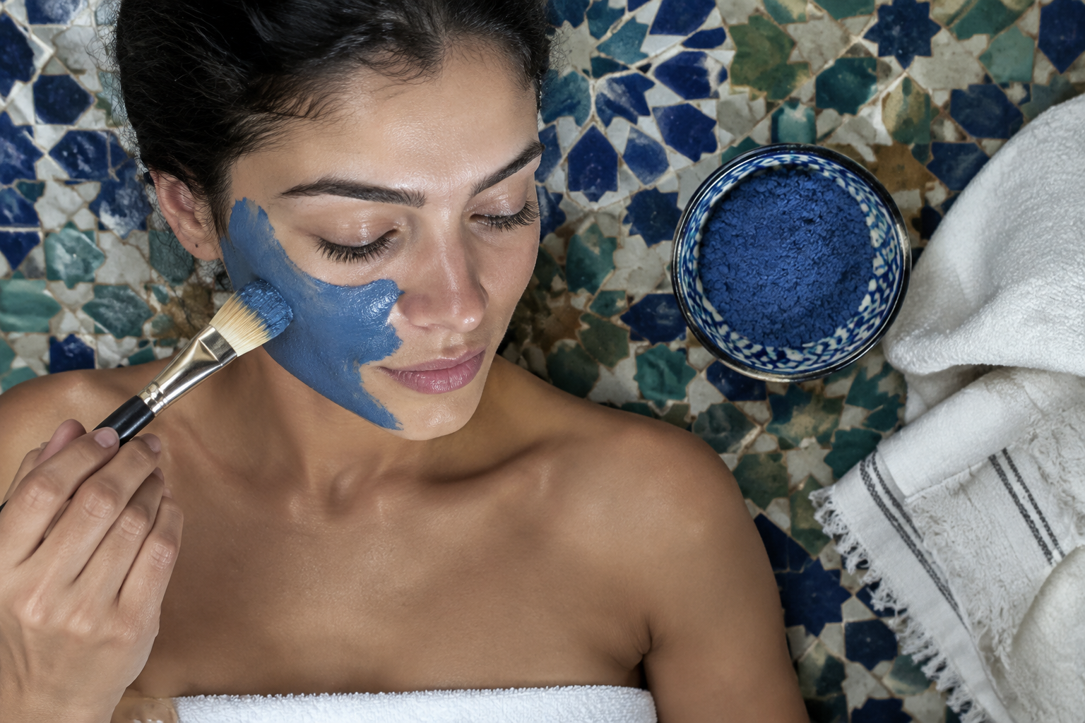 Vrouw brengt een blauw nila gezichtsmasker aan met een kwast, met nila poeder in een kom ernaast